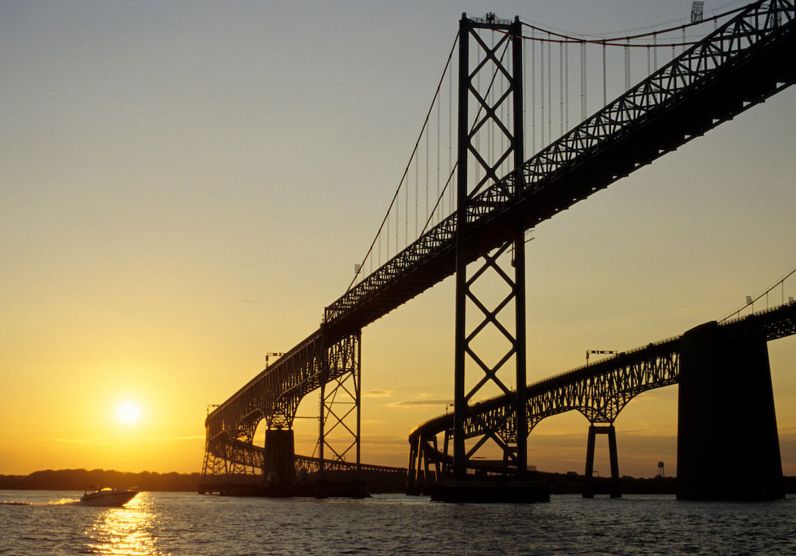 A large span of the Chesapeake Bay Bridge ahead and above with a view of the sun setting and a boat traveling in the water.