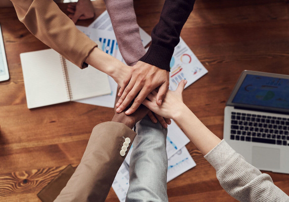 A close up of six individuals laying hands on top of one another, with papers and a laptop below.