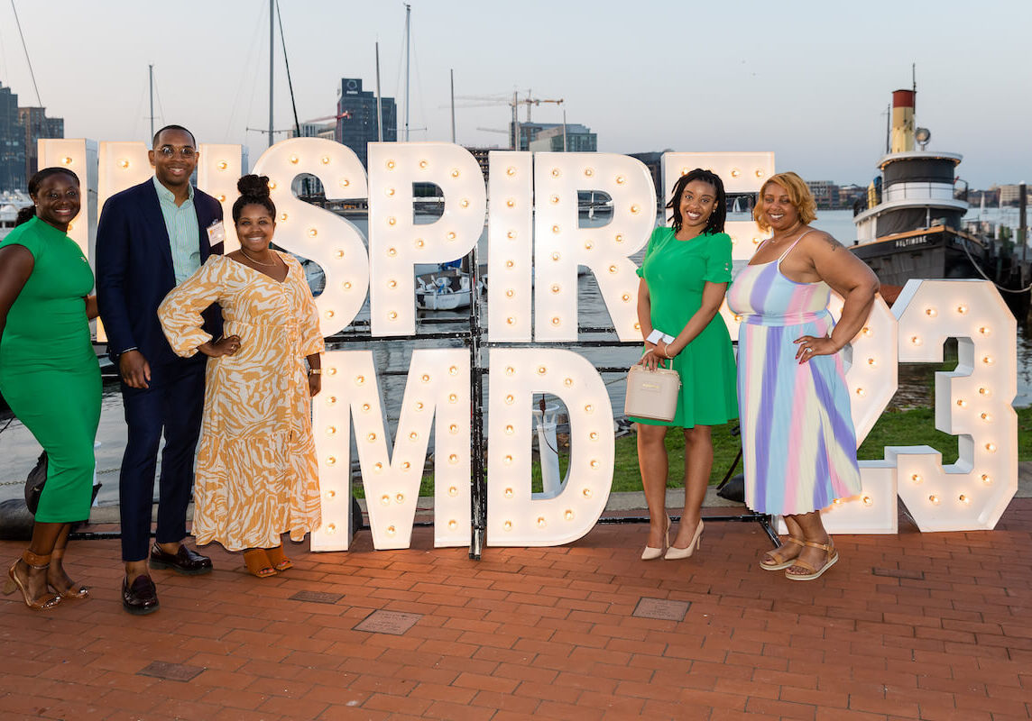 Five people pose in front of a glowing, 3-D Inspire MD sign, with boats and waterfront from Baltimore's Inner Harbor in the background /