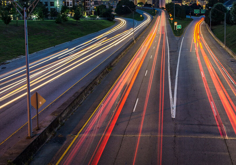 An image at dusk of a freeway with streaks of white head lights on the left and red and yellow tail lights on the right, with views of large city skyscrapers in the distant background.