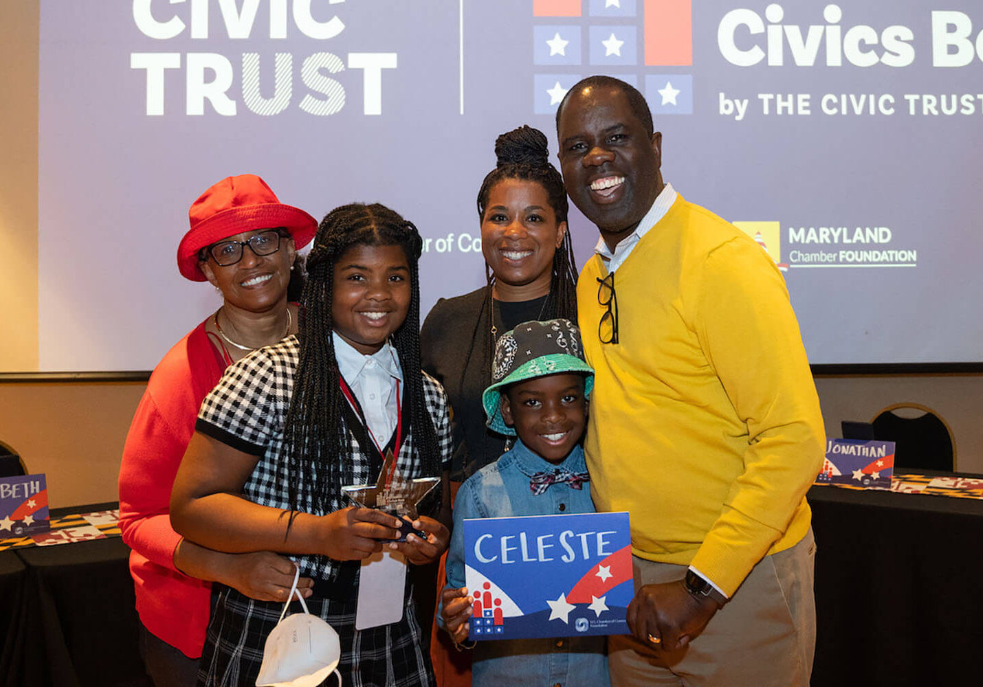 A family with three adults and two children pose and smile while holding a sign with the child's name during the National Civics Bee 2022.