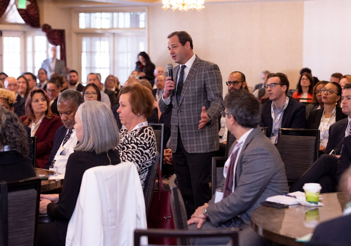 An audience member attending the Maryland Chamber of Commerce Meet the State event stands and uses a microphone to ask questions among an audience of people sitting and listening to remarks being made by members of the Maryland General Assembly.